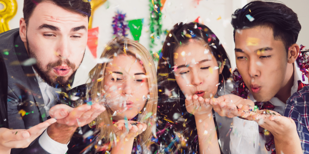 Team of 5 coworkers blow confetti at an in-person office holiday party.