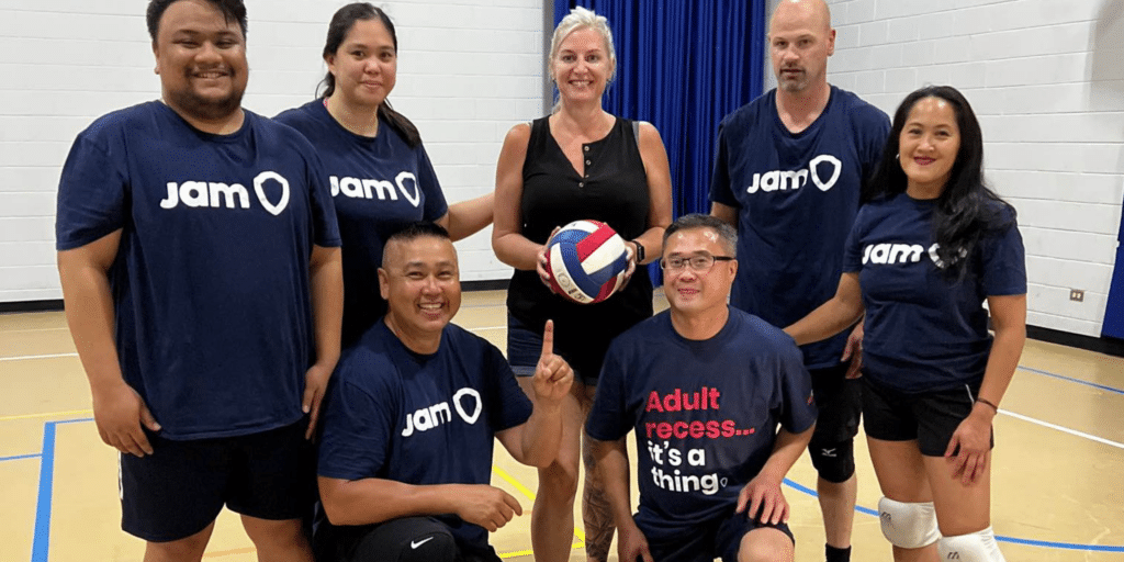 A company sports tournament (court volleyball) to celebrate the holidays. 7 teammates wearing navy blue JAM t-shirts and smiles posing for a team photo with a red, blue and white volleyball. They are in a gym. 